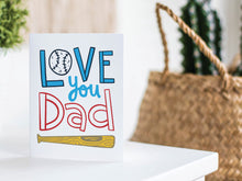 Load image into Gallery viewer, A greeting card is featured on a white tabletop with a white planter in the background with a green plant. There’s a woven basket in the background with a cactus inside. The card features the words “Love You Dad” with an illustrated baseball as the “O” of love and a baseball bat featured at the bottom of the words. 