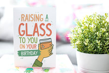 Load image into Gallery viewer, A greeting card is on a table top with a gift in pink wrapping paper. Next to the gift is a white plant pot with a green plant. The card features the words Raising a glass to you on your birthday” with an illustrated hand raising a glass.