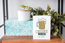 Load image into Gallery viewer, A greeting card is on a table top with a present in blue wrapping paper in the background. On top of the present is a candle and some greenery from a plant too. The card features the words “Cheers to You! Happy Birthday!” with an illustrated beer mug.