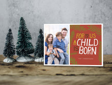 Load image into Gallery viewer, A photo of a one-sided Christmas card showing the front of the card standing up with three small Christmas trees next to it. The photo card features one photo on the left side and on the right side the design is featured with a red background with the words “For to us a child is born, The Thompsons” with green illustrated leaves around the words.