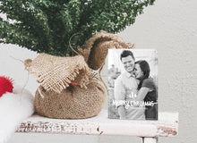 Load image into Gallery viewer, A photo of a one-sided Christmas card showing the front of the card standing up next to the bottom of a small Christmas tree with the base wrapped in burlap. The photo card features one photo with the words “Merry Christmas” with illustrated pine trees above the words. There’s a family name under the words which you can edit with your name.