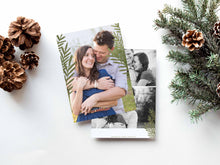 Load image into Gallery viewer, A photo of a double-sided Christmas card showing the front and back of the card laying on a white surface. Around the two sides of the card are pine cones and pine needles. The front of the card features a photo and the words “Comfort in Joy, The Thompsons” in white over the photo. The back of the card features two photos.