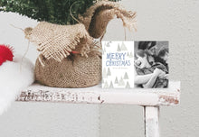 Load image into Gallery viewer, A photo of a one-sided Christmas card showing the front of the card standing up next to the bottom of a small Christmas tree with the base wrapped in burlap. The card features a photo on the right side and to the left side are the words “Merry Christmas” with a family name below it that you can edit. Around the words are whimsical illustrated pine trees.