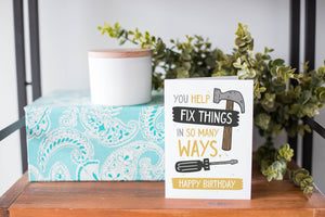 A greeting card is on a table top with a present in blue wrapping paper in the background. On top of the present is a candle and some greenery from a plant too. The card features the words “You help fix things in so many ways. Happy Birthday” featuring an illustrated hammer.