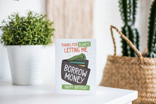 Load image into Gallery viewer, A greeting card is featured on a white tabletop with a white planter in the background with a green plant. There’s a woven basket in the background with a cactus inside. The card features the words “Thanks for always letting me borrow money, happy birthday!” with an illustrated wallet. 