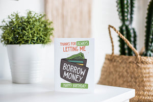 A greeting card is featured on a white tabletop with a white planter in the background with a green plant. There’s a woven basket in the background with a cactus inside. The card features the words “Thanks for always letting me borrow money, happy birthday!” with an illustrated wallet. 