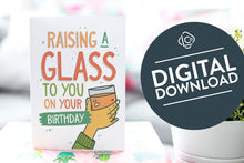 Load image into Gallery viewer, A greeting card is on a table top with a gift in pink wrapping paper. Next to the gift is a white plant pot with a green plant. The card features the words Raising a glass to you on your birthday” with an illustrated hand raising a glass. The words &quot;digital download&quot; are featured in a circle on top of the image. 