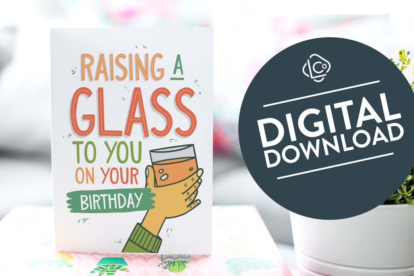 A greeting card is on a table top with a gift in pink wrapping paper. Next to the gift is a white plant pot with a green plant. The card features the words Raising a glass to you on your birthday” with an illustrated hand raising a glass. The words 