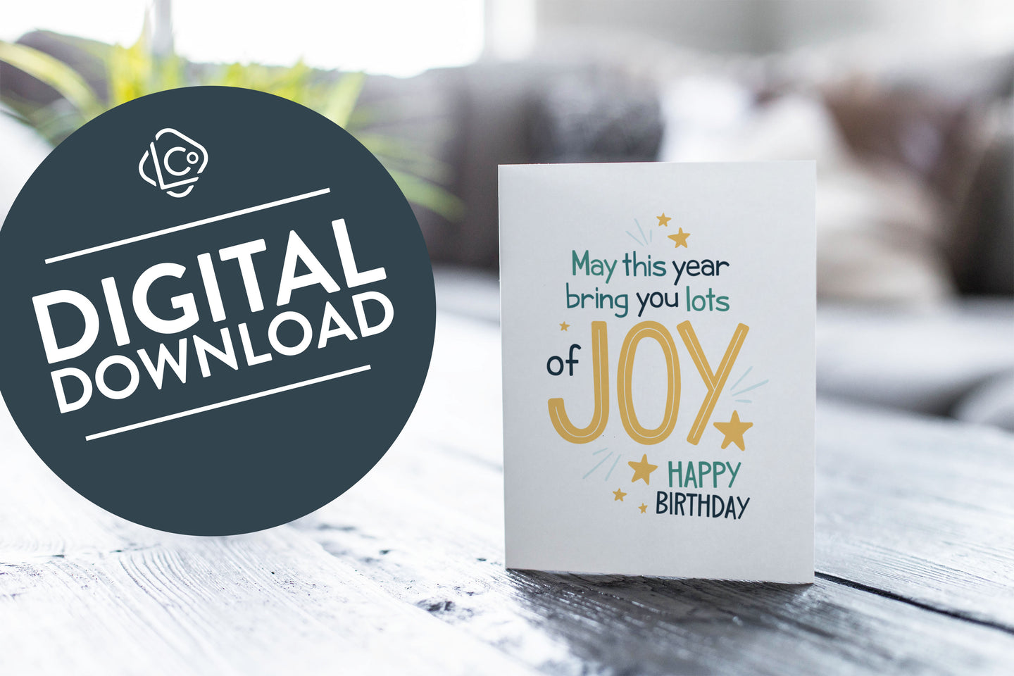 A greeting card is featured on a wood coffee table with a green plant in a white planter in the background. The card features the words “May This Year Bring You Lots of Joy Happy Birthday.”  The words 