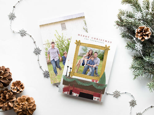 A photo of a double-sided Christmas card showing the front and back of the card laying on a white surface. Around the two sides of the card are pine cones, pine needles and a string of silver snowflake garland. The front of the card features the words “Merry Christmas” with a family name below it that can be edited. A photo is featured in an illustrated frame above an illustrated fireplace. The back of the card features one photo.