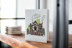 A card on a wood tabletop with an object in the background that is out of focus. The card features an illustrated English village covered in snow with pine trees and smoke coming from the chimneys. 