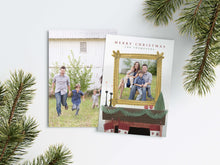 Load image into Gallery viewer, A photo of a Christmas card showing the front and back of the card laying on a white surface. Around the two sides of the card are pine needles. The front of the card features the words “Merry Christmas” with a family name below it that can be edited. A photo is featured in an illustrated frame above an illustrated fireplace. The back of the card features one photo.