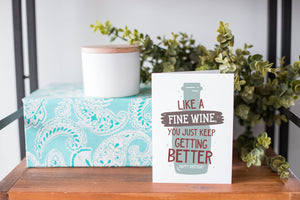 A greeting card is on a table top with a present in blue wrapping paper in the background. On top of the present is a candle and some greenery from a plant too. The card features the words "Like a fine wine, you keep getting better, Happy Birthday!” with an illustration of a wine bottle behind the words.