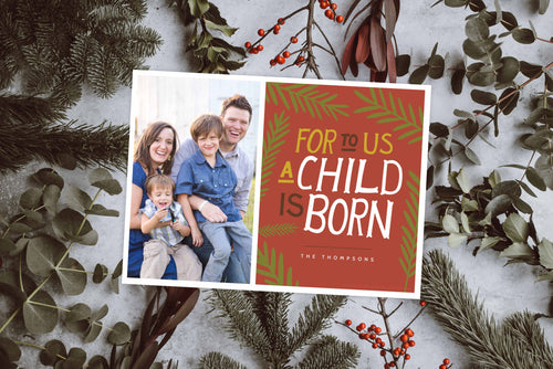A photo of a one-sided Christmas photo card showing the front of the card. The photo card is laying on top of a white surface covered in pine needles, eucalyptus leaves, and branches with red berries.The photo card features one photo on the left side and on the right side the design is featured with a red background with the words “For to us a child is born, The Thompsons” with green illustrated leaves around the words. 