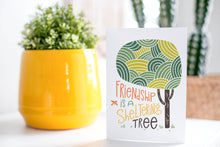 Load image into Gallery viewer, A greeting card is on a table top with a yellow plant pot and a green plant inside. The card features the words “Friendship is a sheltering tree” with an illustrated tree.