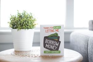 A greeting card laying on a wooden table with some cut wood details. The card features the words “Thanks for always letting me borrow money, happy birthday!” with an illustrated wallet.