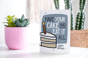  A greeting card featured standing up on a white tabletop with a pink plant pot in the background and some succulents in the pot. There’s a woven basket in the background with a cactus inside. The card features the words “Have Your cake and eat it all, Happy birthday!” with an illustrated piece of cake with a candle on the top.