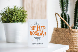 A greeting card is featured on a white tabletop with a white planter in the background with a green plant. There’s a woven basket in the background with a cactus inside. The card features the words “Hip Hip Hooray! Happy Birthday!”