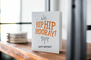 A card on a wood tabletop with an object in the background that is out of focus. The card features the words “Hip Hip Hooray! Happy Birthday!” The words "digital download" are featured in a circle on top of the image.