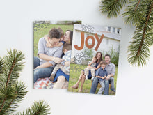 Load image into Gallery viewer, A photo of a Christmas card showing the front and back of the card laying on a white surface. Around the two sides of the card are pine needles. The front of the card features a photo with the word “Joy” in an orange-red color and some gray illustrated modern leaves. The back of the card features one photo.