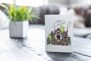 A greeting card featured on a black, wood coffee table. There’s a white planter in the background with a green plant. There’s also a gray sofa in the background with a white pillow. The card features an illustrated English village covered in snow with pine trees and smoke coming from the chimneys.