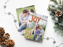 Load image into Gallery viewer, A photo of a double-sided Christmas card showing the front and back of the card laying on a white surface. Around the two sides of the card are pine cones, pine needles and a string of silver snowflake garland. The front of the card features a photo with the word “Joy” in an orange-red color and some gray illustrated modern leaves. The back of the card features one photo.