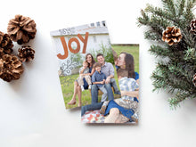 Load image into Gallery viewer, A photo of a double-sided Christmas card showing the front and back of the card laying on a white surface. Around the two sides of the card are pine cones and pine needles. The front of the card features a photo with the word “Joy” in an orange-red color and some gray illustrated modern leaves. The back of the card features one photo.