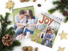 Load image into Gallery viewer, A photo of a two-sided Christmas card showing the front of the card on top of a brown wrapped gift on a white tabletop. Around the gift are pine needles, pinecones and wood star ornaments. The front of the card features a photo with the word “Joy” in an orange-red color and some gray illustrated modern leaves. The back of the card features one photo.