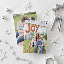 Load image into Gallery viewer, A photo of a double-sided Christmas card showing the front and back of the card laying on a white surface. Around the two sides of the card are surrounded with white snowflake ornaments, silver bells, a silver reindeer, white tea light snowflake candle and silver star garland. The front of the card features a photo with the word “Joy” in an orange-red color and some gray illustrated modern leaves. The back of the card features one photo.