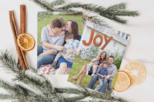 A photo of a double-sided Christmas card showing the front and back of the card laying on a white surface. Around the two sides of the card are pine needles, cinnamon sticks and dried oranges. The front of the card features a photo with the word “Joy” in an orange-red color and some gray illustrated modern leaves. The back of the card features one photo. 