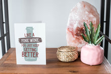 Load image into Gallery viewer, A card on a wood tabletop and on the right side of the card is a woven basket, a pink plant pot with a cactus in it and a pink crystal rock. The card features the words &quot;Like a fine wine, you keep getting better, Happy Birthday!” with an illustration of a wine bottle behind the words.