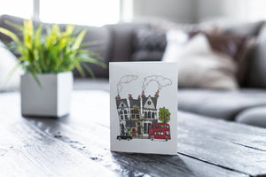 A greeting card featured on a black, wood coffee table. There’s a white planter in the background with a green plant. There’s also a gray sofa in the background with a white pillow. The card features a design with illustrated London houses, a black taxi cab and a red double decker bus.