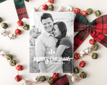 Load image into Gallery viewer, A photo of a one-sided Christmas card showing the front of the card laying on white table top. The card is surrounded by plaid red and green ribbon and red, green and white bells. The photo card features one photo with the words “Merry Christmas” with illustrated pine trees above the words. There’s a family name under the words which you can edit with your name.