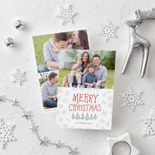 Load image into Gallery viewer, A photo of a double-sided Christmas card showing the front and back of the card laying on a white surface. Around the two sides of the card are surrounded with Christmas items. The front of the card features a photo on the top half of the card with the words “Merry Christmas” in a fun font with illustrated pine trees below the words. Below the pine trees you can customise with your family name. The back of the card features two photos along with illustrated snowflakes and pine trees.