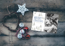 Load image into Gallery viewer, A photo of a one-sided Christmas card showing the front and back of the card laying on a wood surface. To the left of the cards are some ornaments. The card features a photo on the right side and to the left side are the words “Merry Christmas” with a family name below it that you can edit. Around the words are whimsical illustrated pine trees.