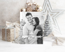 Load image into Gallery viewer, A photo of a one-sided Christmas card showing the front of the card standing up with Christmas items behind the card. There’s a sparkly star, glitter wrapped gift, ribbon and ornaments with a small silver tree around the photo card. The photo card features one photo with the words “Merry Christmas” with illustrated pine trees above the words. There’s a family name under the words which you can edit with your name.