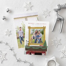 Load image into Gallery viewer, A photo of a double-sided Christmas card showing the front and back of the card laying on a white surface. Around the two sides of the card are surrounded with white snowflake ornaments, silver bells, a silver reindeer, white tea light snowflake candle and silver star garland. The front of the card features the words “Merry Christmas” with a family name below it that can be edited. A photo is featured in an illustrated frame above an illustrated fireplace. The back of the card features one photo.