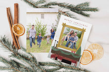 Load image into Gallery viewer, A photo of a double-sided Christmas card showing the front and back of the card laying on a white surface. Around the two sides of the card are pine needles, cinnamon sticks and dried oranges. The front of the card features the words “Merry Christmas” with a family name below it that can be edited. A photo is featured in an illustrated frame above an illustrated fireplace. The back of the card features one photo.
