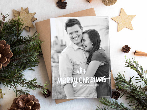 A photo of a one-sided Christmas card showing the front of the card on top of a brown wrapped gift on a white tabletop. Around the gift are pine needles, pinecones and wood star ornaments. The photo card features one photo with the words “Merry Christmas” with illustrated pine trees above the words. There’s a family name under the words which you can edit with your name. 