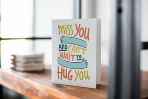 A card on a wood tabletop with an object in the background that is out of focus. The card features the words “Miss you and can’t wait to hug you.”