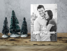 Load image into Gallery viewer, A photo of a one-sided Christmas card showing the front of the card standing up with three small Christmas trees next to it. The photo card features one photo with the words “Merry Christmas” with illustrated pine trees above the words. There’s a family name under the words which you can edit with your name.