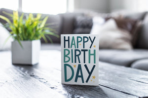 A greeting card is featured on a wood coffee table with a green plant in a white planter in the background. The card features the words “Happy birthday” with blue letters featured on a white background. 