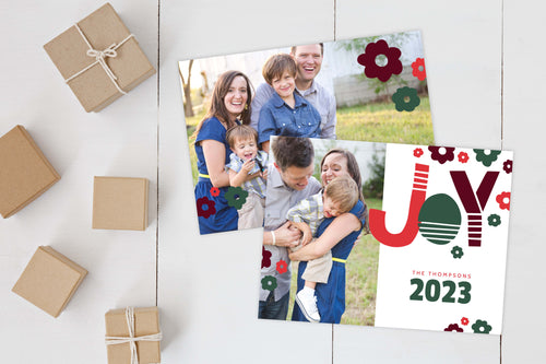 A photo of a Christmas card showing the front and back of the card laying on a white surface. Brown wrapped small gifts are to the left of the cards. The front of the card features a photo to the left side and to the right side the word “Joy” is featured in a fun, modern font with modern illustrated flowers around it. Below the word is the option to put your family name and the year 2023. The back of the card features two photos with the modern illustrated flowers. 
