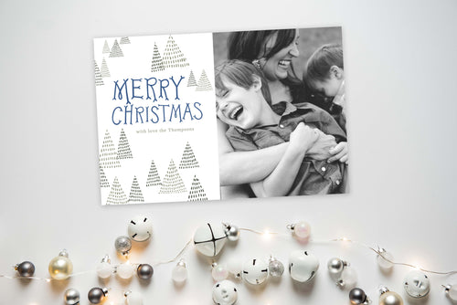 A photo of a one-sided Christmas card showing the front and back of the card laying on a white surface. To the bottom of the cards are silver and white small ornaments. The card features a photo on the right side and to the left side are the words “Merry Christmas” with a family name below it that you can edit. Around the words are whimsical illustrated pine trees. 