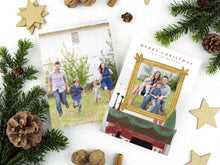 Load image into Gallery viewer, A photo of a two-sided Christmas card showing the front of the card on top of a brown wrapped gift on a white tabletop. Around the gift are pine needles, pinecones and wood star ornaments. The front of the card features the words “Merry Christmas” with a family name below it that can be edited. A photo is featured in an illustrated frame above an illustrated fireplace. The back of the card features one photo.
