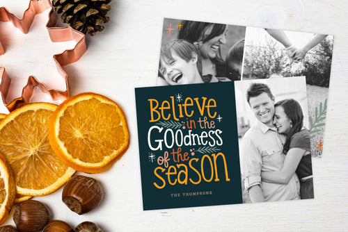 A photo of a Christmas card showing the front and back of the card laying on a white surface. Left of the card is a cookie cutter, pinecone, nuts and dried oranges. The front of the card features a photo and the words “Believe in the goodness of the season, The Thompsons” to the left of the photo. The back of the card features two photos. 