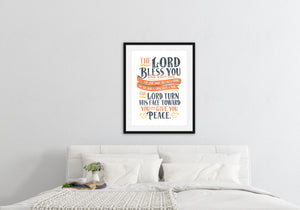 Artwork is featured in a black frame on a white wall above a bed with white linens. The artwork features hand drawn lettering of the Bible verse Numbers 6:24-26 reading "The Lord bless you and keep you. The Lord make his face to shine on you and be gracious to you. The Lord turn his face toward you and give you peace."