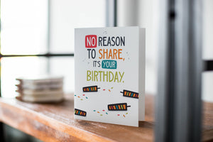 A card on a wood tabletop with an object in the background that is out of focus. The card features the words “No reason to share it’s your birthday!”