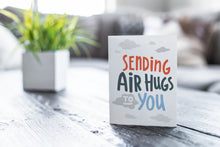 Load image into Gallery viewer, A greeting card featured on a black, wood coffee table. There’s a white planter in the background with a green plant. There’s also a gray sofa in the background with a white pillow. The card features the words “Sending air hugs to you.”