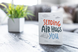 A greeting card featured on a black, wood coffee table. There’s a white planter in the background with a green plant. There’s also a gray sofa in the background with a white pillow. The card features the words “Sending air hugs to you.”